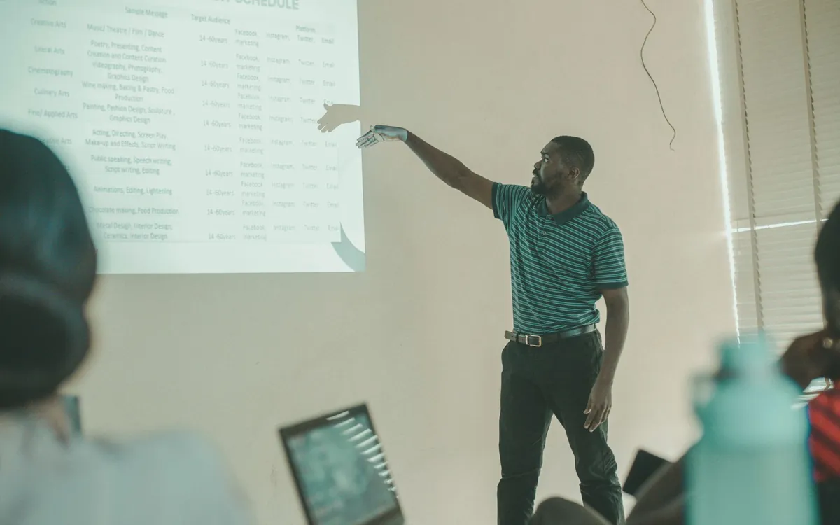 In a classroom, a teacher points at a table projected on the wall. In the foreground, seen from behind, students and an open laptop.