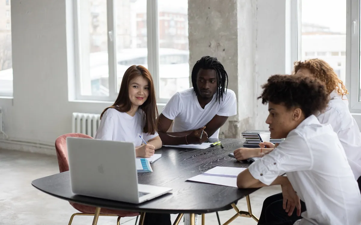 Four students around a round table, in a bright room, talking in front of a laptop and sheets of notes.